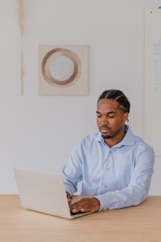 A professional man with braided hair working on a laptop in a modern office setting.