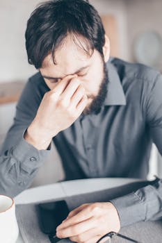 Bearded man looking stressed and tired, sitting at a desk with paperwork.