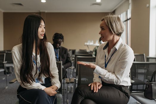 Two women in business attire discuss topics at a professional conference, seated indoors.